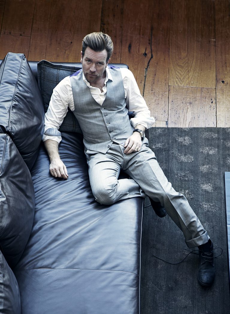Ewan McGregor poses on a blue leather couch, wearing a white button-down shirt, grey vest, and grey pants in a high-angle shot.