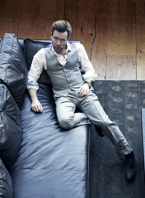 Ewan McGregor poses on a blue leather couch, wearing a white button-down shirt, grey vest, and grey pants in a high-angle shot.