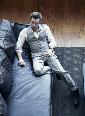 Ewan McGregor poses on a blue leather couch, wearing a white button-down shirt, grey vest, and grey pants in a high-angle shot.