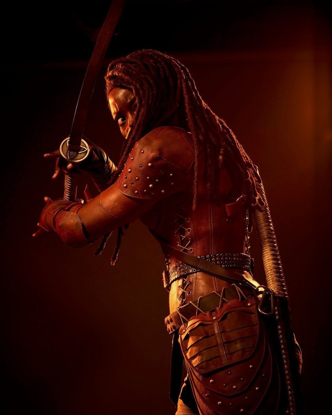 Adult female warrior in red studded leather armor holding a katana, standing in a low-lit photo studio with an intense expression.