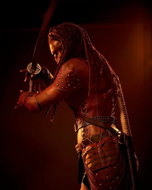 Adult female warrior in red studded leather armor holding a katana, standing in a low-lit photo studio with an intense expression.