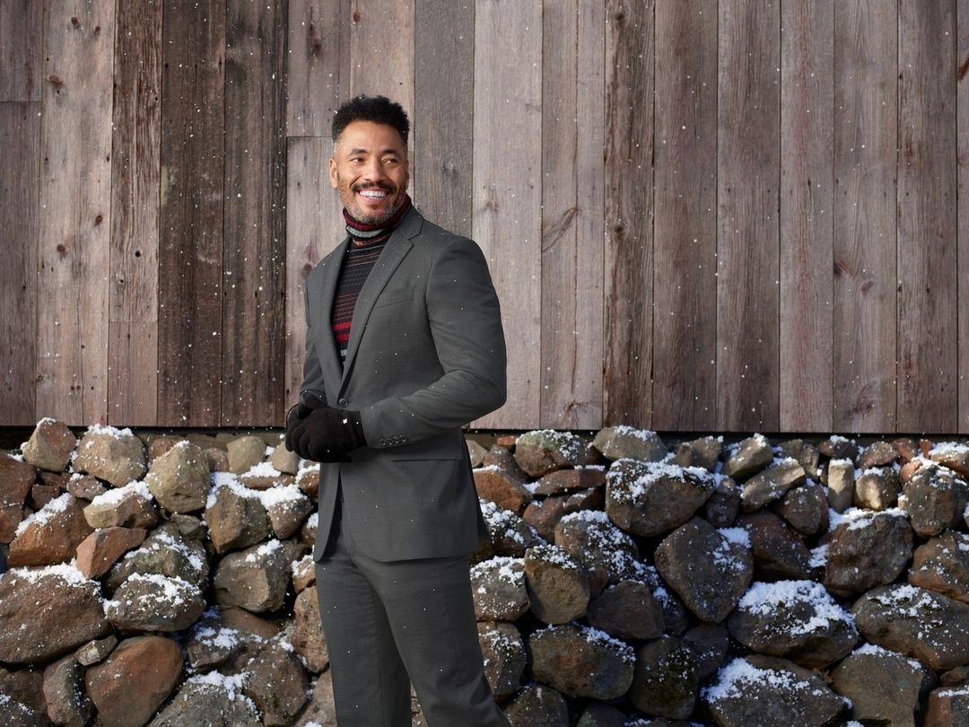 Man in a gray suit stands in front of a wooden wall and a rocky, snowy ground, smiling and looking to the side in an outdoor setting.