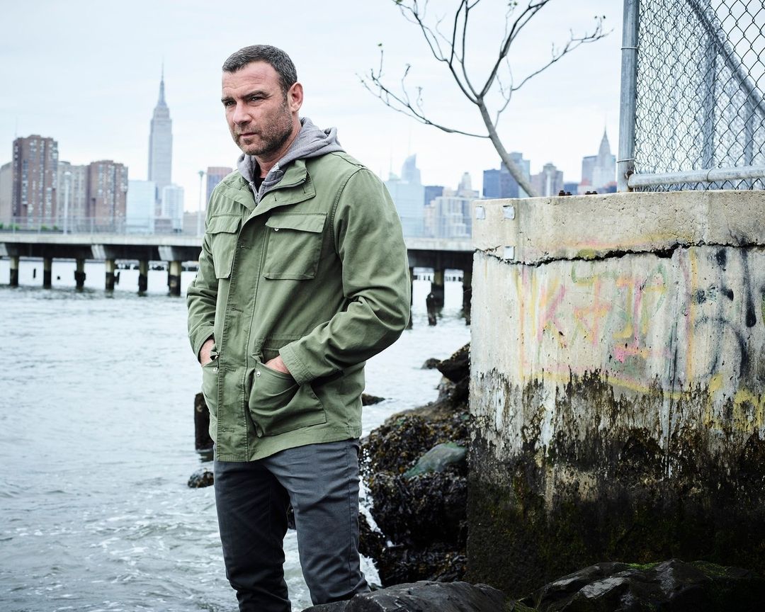 A man in a green jacket stands near the water's edge with a city skyline featuring the Empire State Building in the background on a cloudy day.