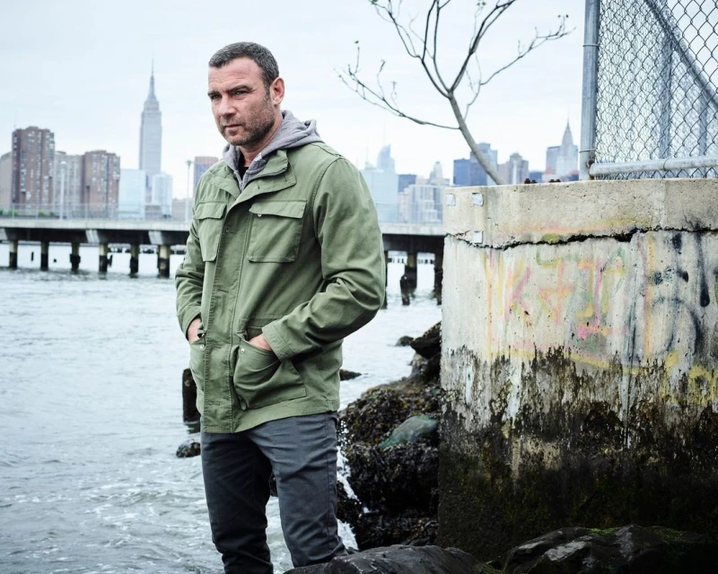 A man in a green jacket stands near the water's edge with a city skyline featuring the Empire State Building in the background on a cloudy day.