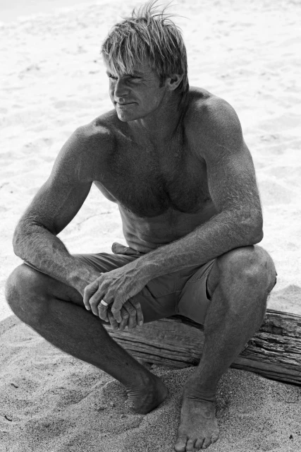 Black and white portrait of a muscular adult man sitting on a log on a beach, wearing shorts and looking to the left.