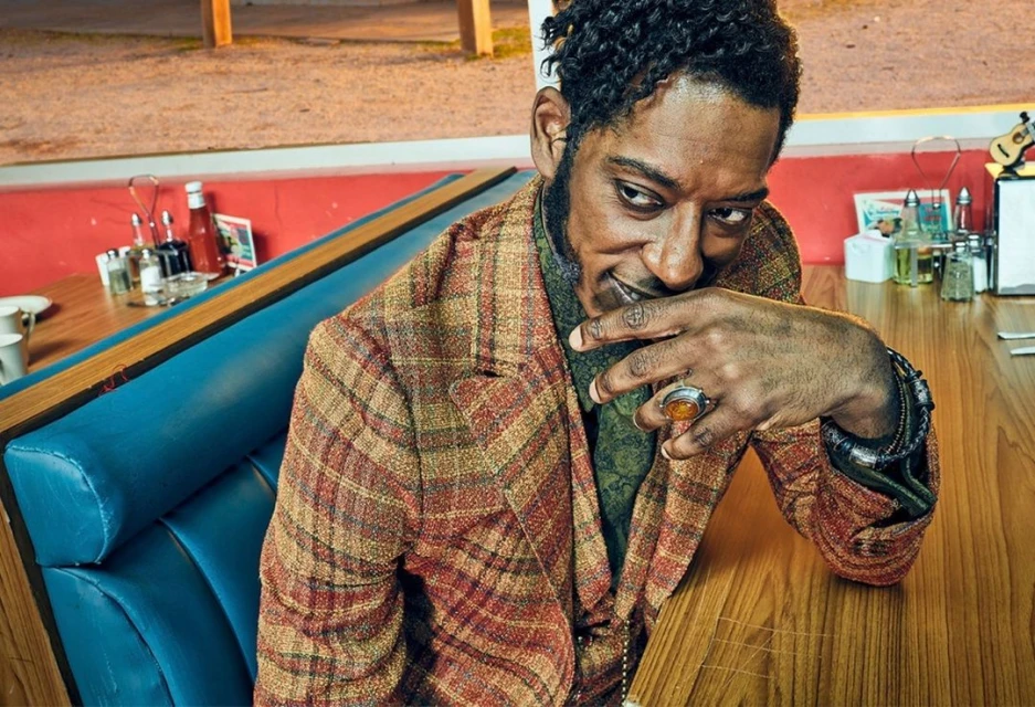 Adult male wearing a plaid blazer and bracelets, seated at a diner booth. The setting includes a table and diner condiments.