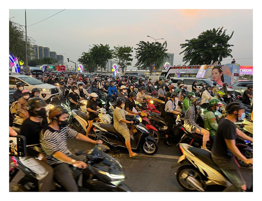Busy urban scene with a large group of people on motorcycles and scooters in a city street with buildings in the background at dusk.