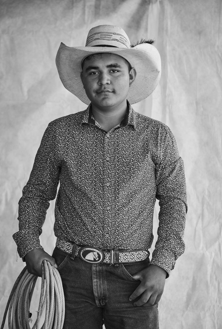 Black and white portrait of a young adult wearing a cowboy hat, patterned shirt, jeans, and belt with horse buckle, holding a rope.