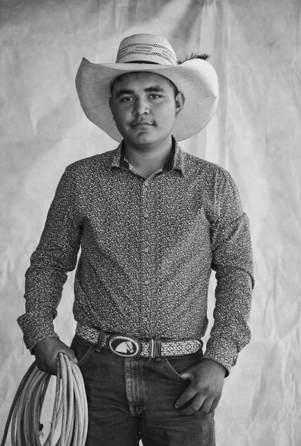 Black and white portrait of a young adult wearing a cowboy hat, patterned shirt, jeans, and belt with horse buckle, holding a rope.