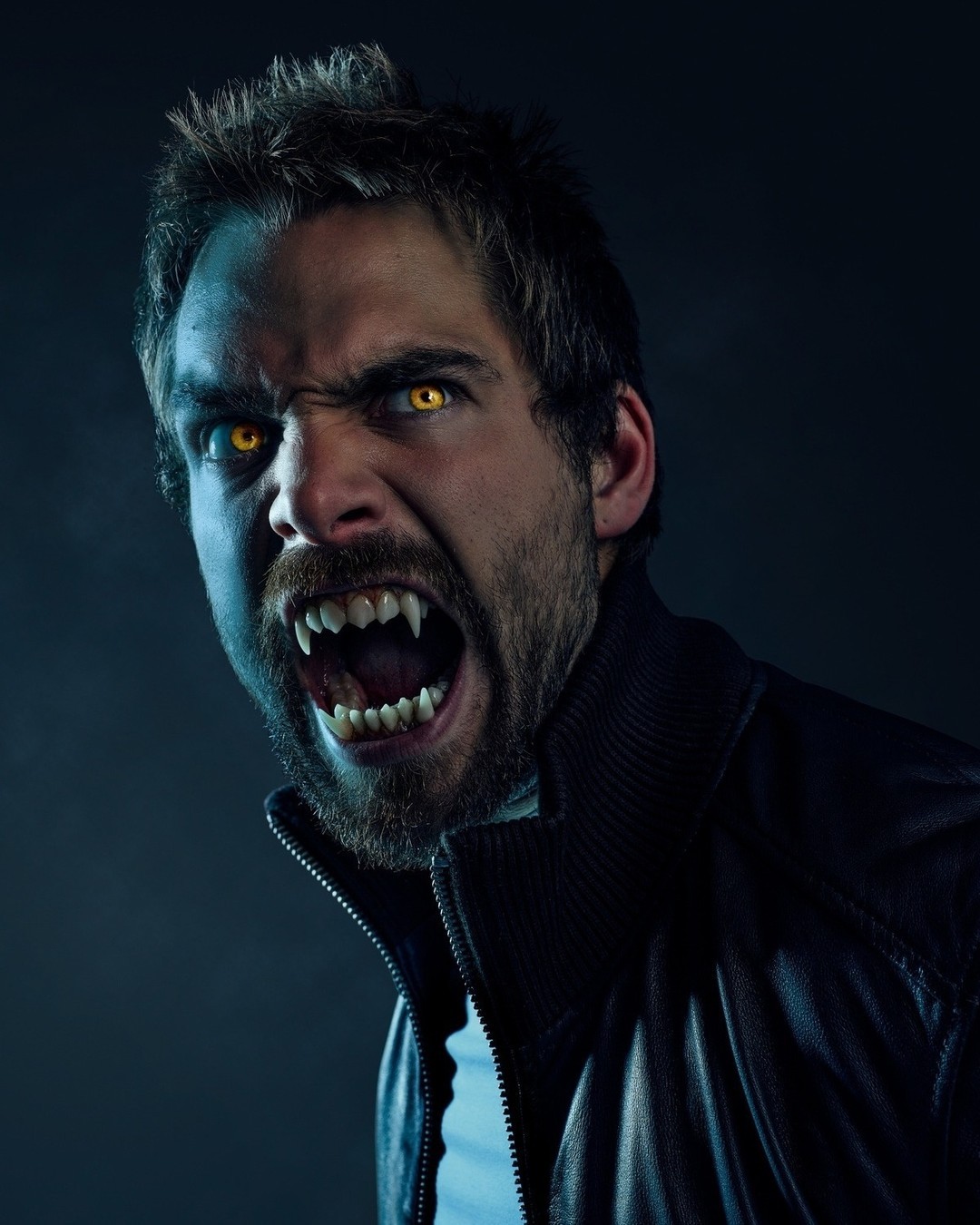 Close-up of a Caucasian male with a beard, bared fangs, and yellow eyes, wearing a black leather jacket against a dark blue backdrop.