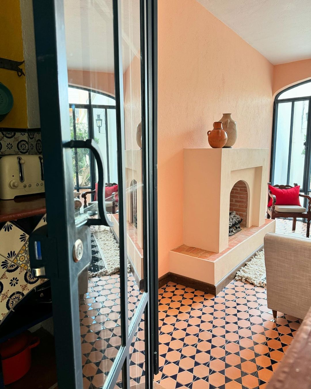 Interior view through glass doors of a room with terracotta tile flooring, a faux fireplace with vases, and seating in natural light.