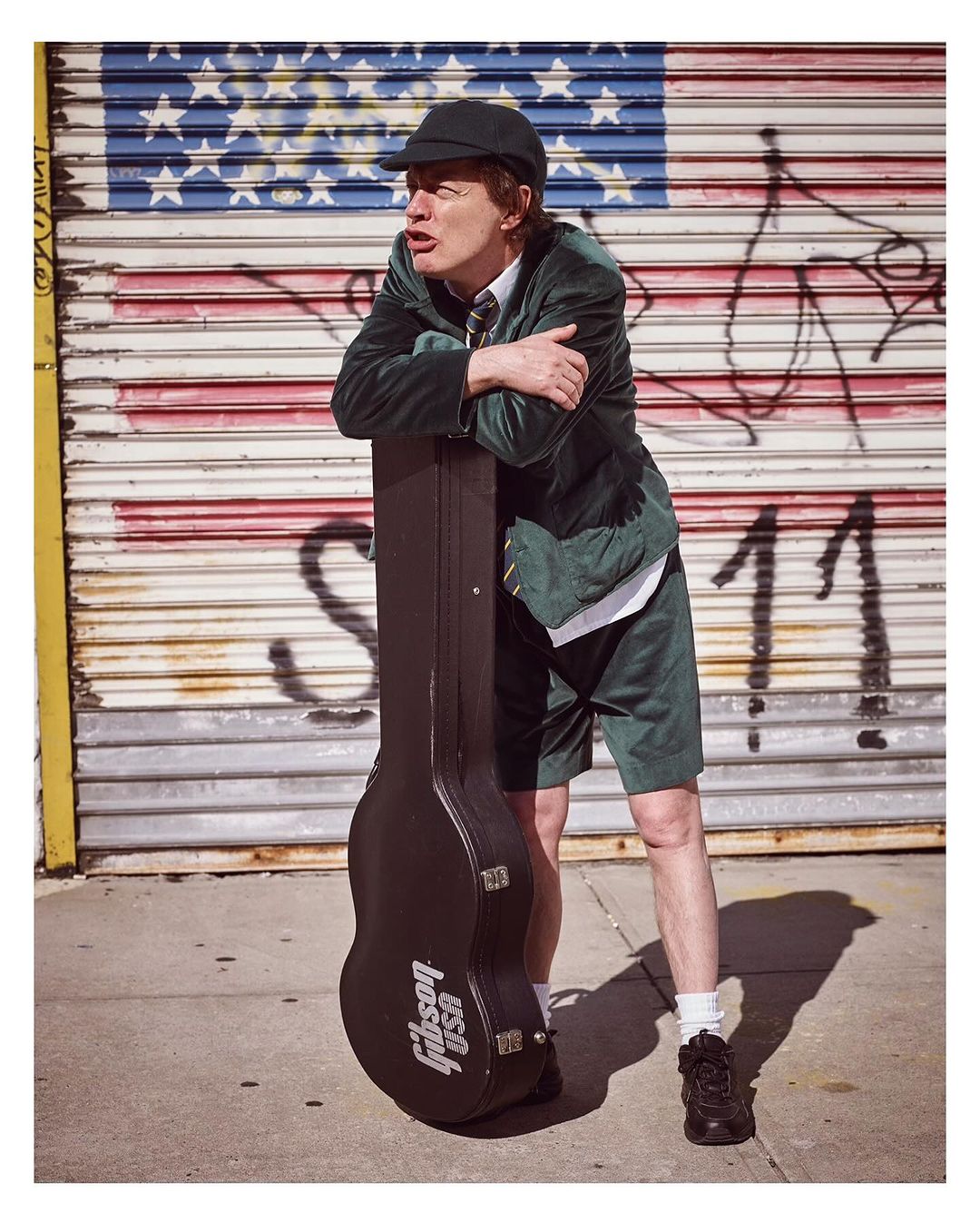 Brian Johnson of AC/DC poses with a guitar case against an American flag painted on a corrugated metal wall on a city street.