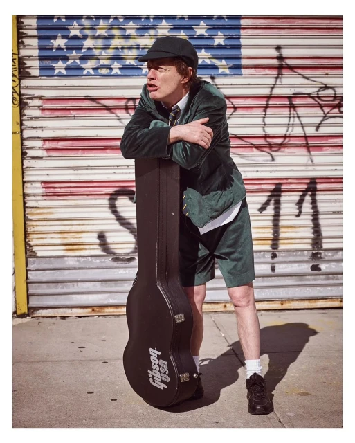 Brian Johnson of AC/DC poses with a guitar case against an American flag painted on a corrugated metal wall on a city street.