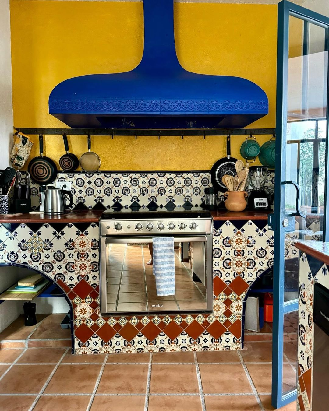 Indoor kitchen with a blue range hood above a stainless-steel stove, decorative tile backsplash, terracotta tiled floor, and yellow wall.