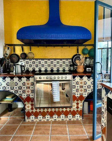 Indoor kitchen with a blue range hood above a stainless-steel stove, decorative tile backsplash, terracotta tiled floor, and yellow wall.