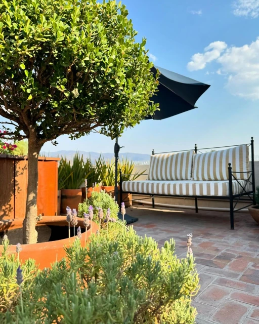 Outdoor seating area on a brick patio featuring a bench with striped cushions, potted plants, a shade umbrella, and a mountain view.