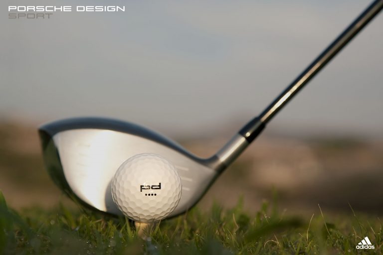 Close-up of a golf ball on a tee with a golf club behind it, outdoors with a blurred green and tan background, and logos visible.