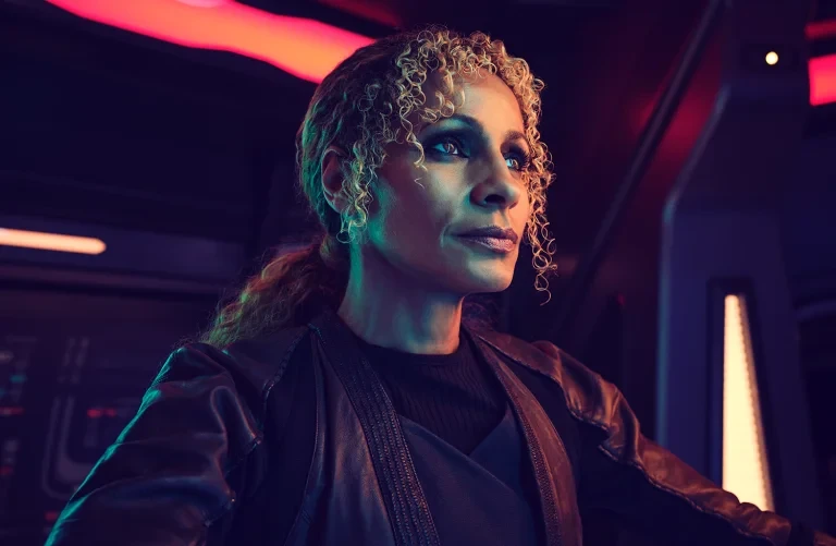 Portrait of a woman with curly hair in a dark spaceship interior with blue and red lighting, looking intently off-camera.