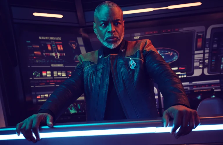 Adult male with light gray hair in a black jacket, standing in front of a computer console with blue lights. Science fiction setting.