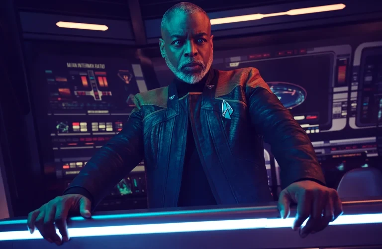 Adult male with light gray hair in a black jacket, standing in front of a computer console with blue lights. Science fiction setting.
