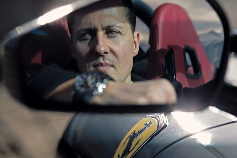 Close-up on an adult male driving a sports car, wearing a watch, with a desert mountain landscape visible in the background.