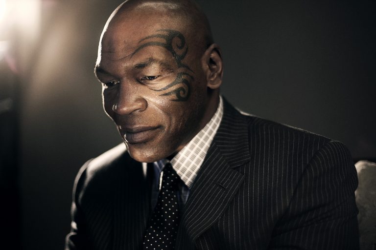 Portrait of an African American adult male with a face tattoo, wearing a pinstripe suit and patterned tie, looking downward with a serious expression.