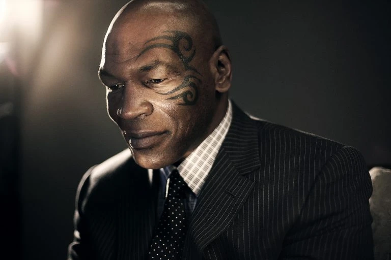 Portrait of an African American adult male with a face tattoo, wearing a pinstripe suit and patterned tie, looking downward with a serious expression.