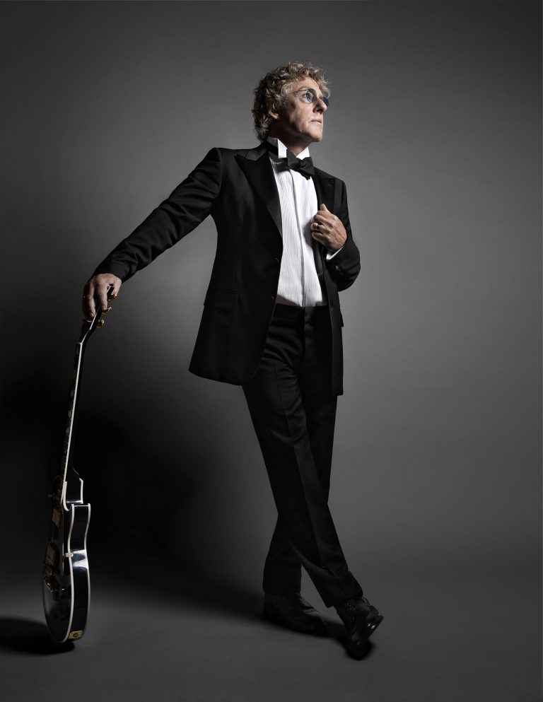 An older man wearing a black tuxedo and bow tie holding an electric guitar against a dark grey background.