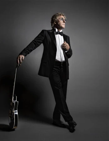An older man wearing a black tuxedo and bow tie holding an electric guitar against a dark grey background.