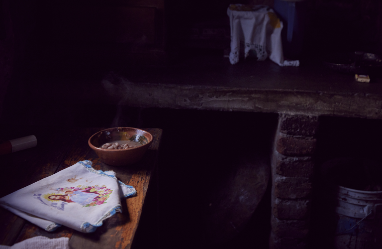 A rustic wooden table in a dimly lit room holds a ceramic bowl of beans and a folded white cloth decorated with an embroidered image.