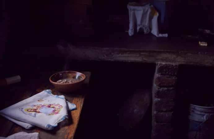 A rustic wooden table in a dimly lit room holds a ceramic bowl of beans and a folded white cloth decorated with an embroidered image.