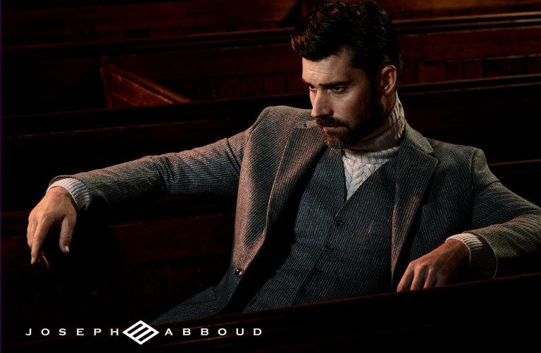 Stylish adult male in a grey suit, sitting inside a building with dark wood benches, looking to the side with a neutral expression.
