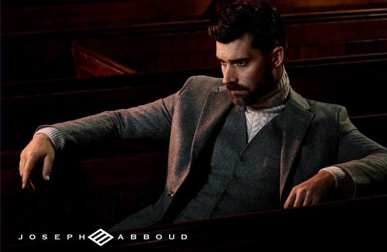 Stylish adult male in a grey suit, sitting inside a building with dark wood benches, looking to the side with a neutral expression.