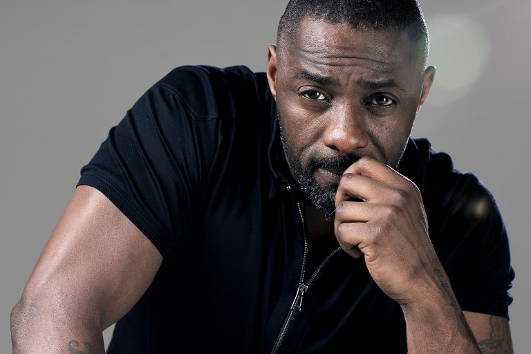 Portrait of Idris Elba wearing a black zipped top with hand near mouth against a neutral grey studio background