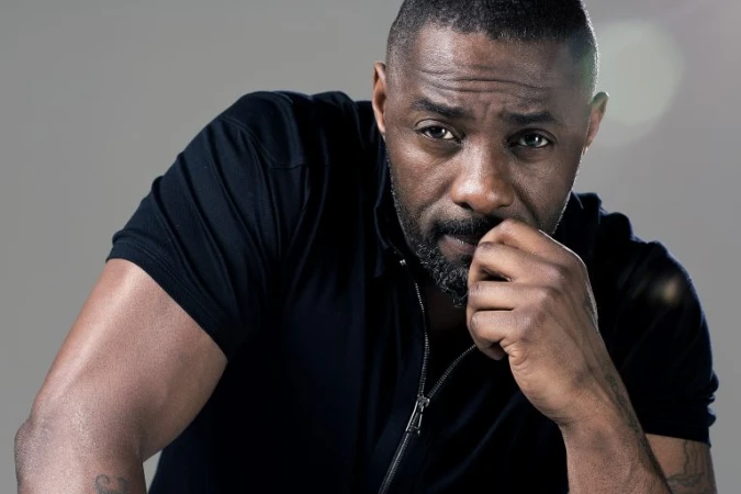 Portrait of Idris Elba wearing a black zipped top with hand near mouth against a neutral grey studio background