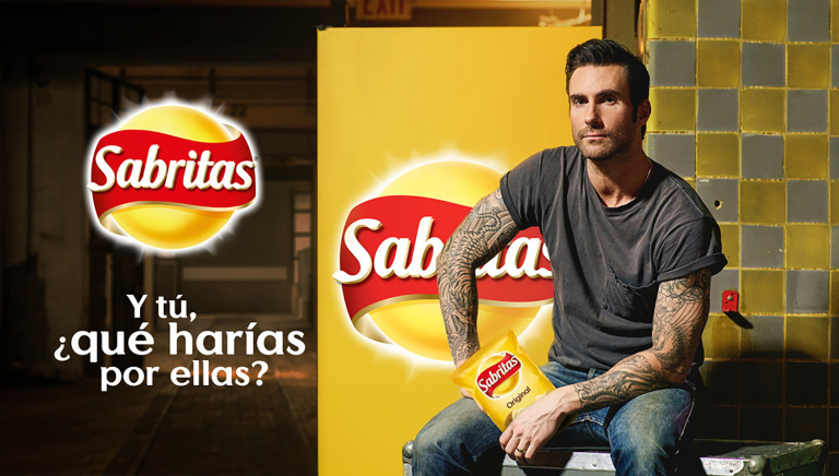Adam Levine poses with a bag of Sabritas Original chips in a marketing shot. He sits indoors against a graphic backdrop.