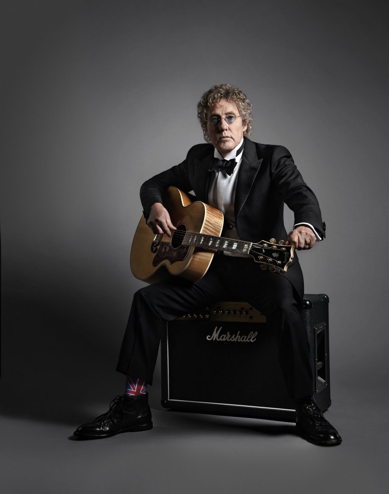 Portrait of an adult male seated holding an acoustic guitar, wearing a tuxedo and dress shoes in a photo studio environment.