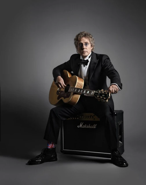 Portrait of an adult male seated holding an acoustic guitar, wearing a tuxedo and dress shoes in a photo studio environment.