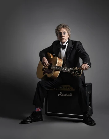 Portrait of an adult male seated holding an acoustic guitar, wearing a tuxedo and dress shoes in a photo studio environment.
