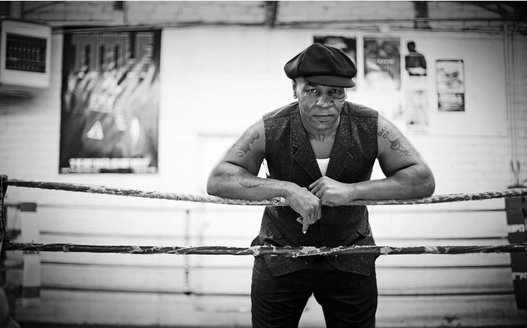 Monochrome portrait of a man in a cap leaning against a boxing ring, wearing a vest, with boxing posters in the background.