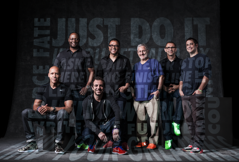 Group portrait of seven men of mixed ages and ethnicities wearing casual clothing and sneakers in front of a gray background with text.