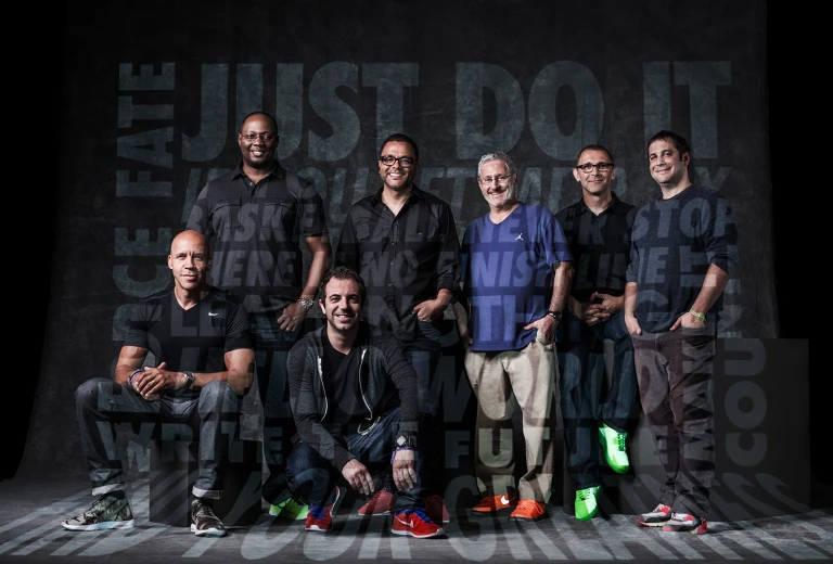 Group portrait of seven men of mixed ages and ethnicities wearing casual clothing and sneakers in front of a gray background with text.