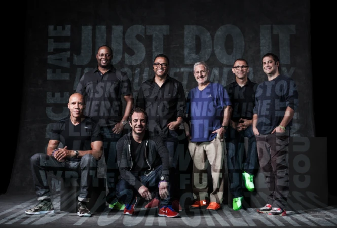 Group portrait of seven men of mixed ages and ethnicities wearing casual clothing and sneakers in front of a gray background with text.