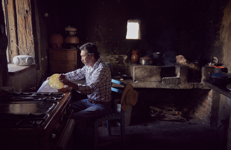 Adult male in a rustic kitchen prepares food. The interior features a wood-burning stove, cabinets, and dim lighting, creating a vintage setting.