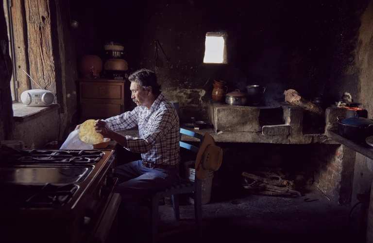 Adult male in a rustic kitchen prepares food. The interior features a wood-burning stove, cabinets, and dim lighting, creating a vintage setting.