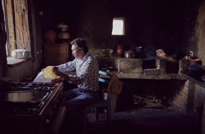 Adult male in a rustic kitchen prepares food. The interior features a wood-burning stove, cabinets, and dim lighting, creating a vintage setting.