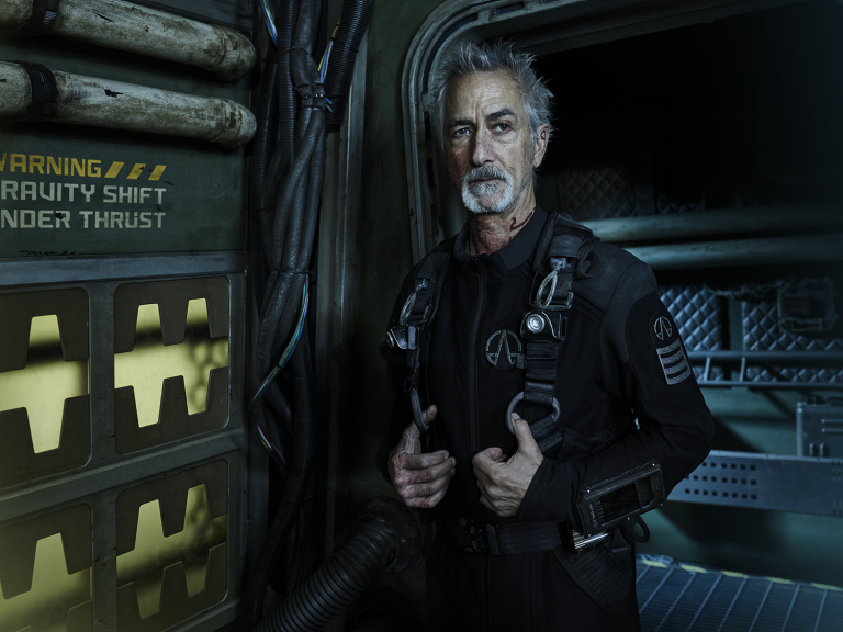 A man with grey hair and a goatee in a dark jumpsuit poses in a dimly lit, metallic corridor with warning signs in a science fiction setting.