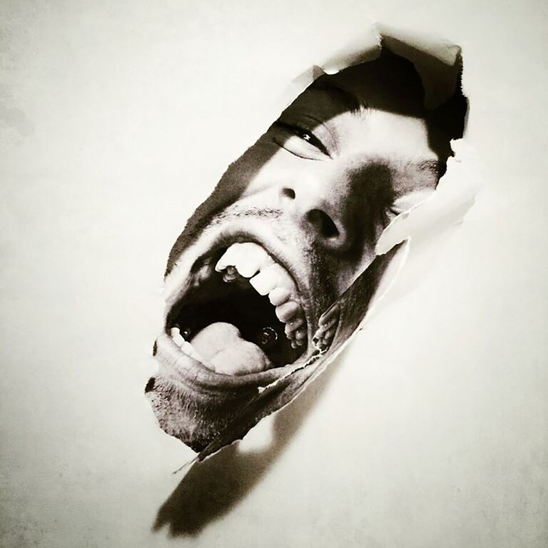 Monochrome portrait of a person's face emerging from a torn paper background, mouth open in a shout, teeth visible, intense expression.