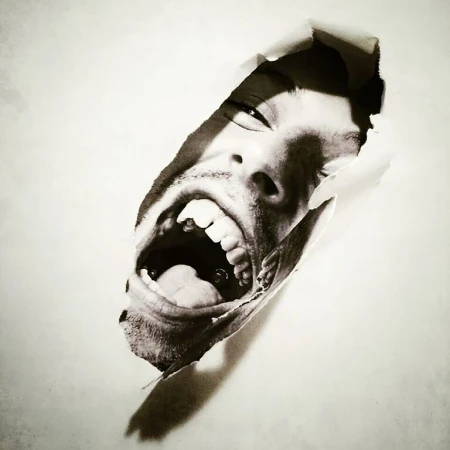 Monochrome portrait of a person's face emerging from a torn paper background, mouth open in a shout, teeth visible, intense expression.