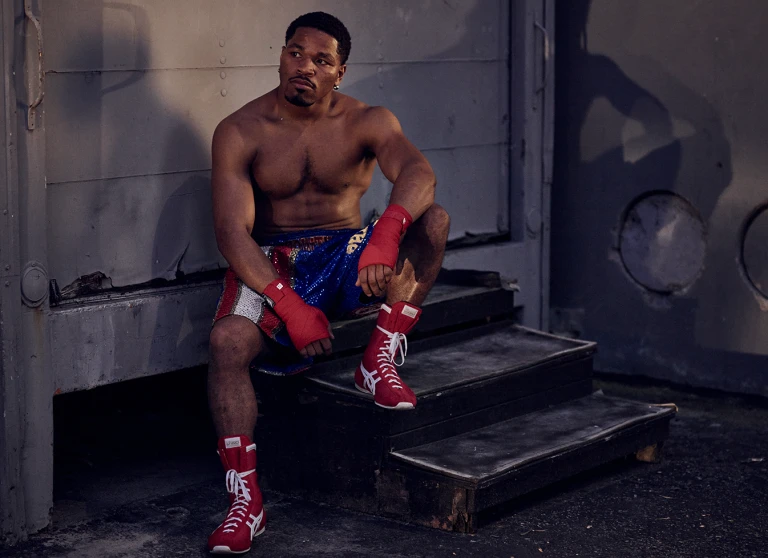 Muscular shirtless man in boxing shorts and red boots sits on gray steps, looking to the side, hands wrapped in red cloth.
